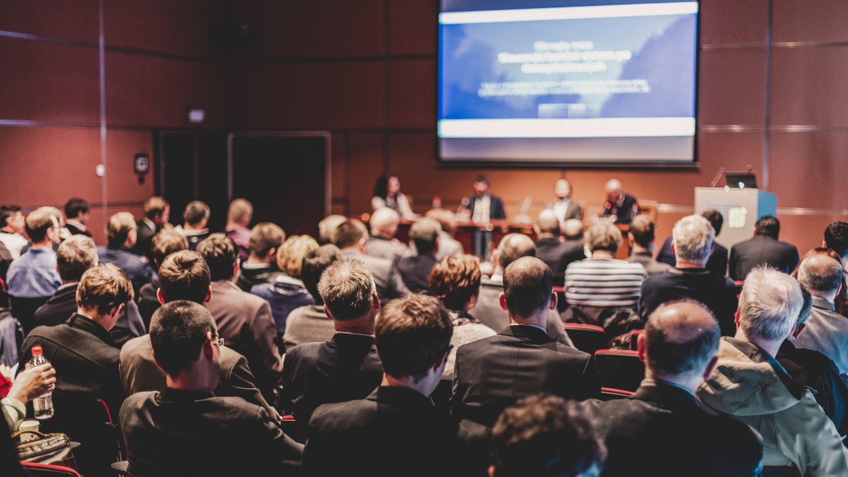 "A large audience seated in a conference hall watching a panel of speakers on stage with a presentation screen, illustrating the scale of human connection that AI now optimizes through data-driven event marketing and personalized attendee experiences.