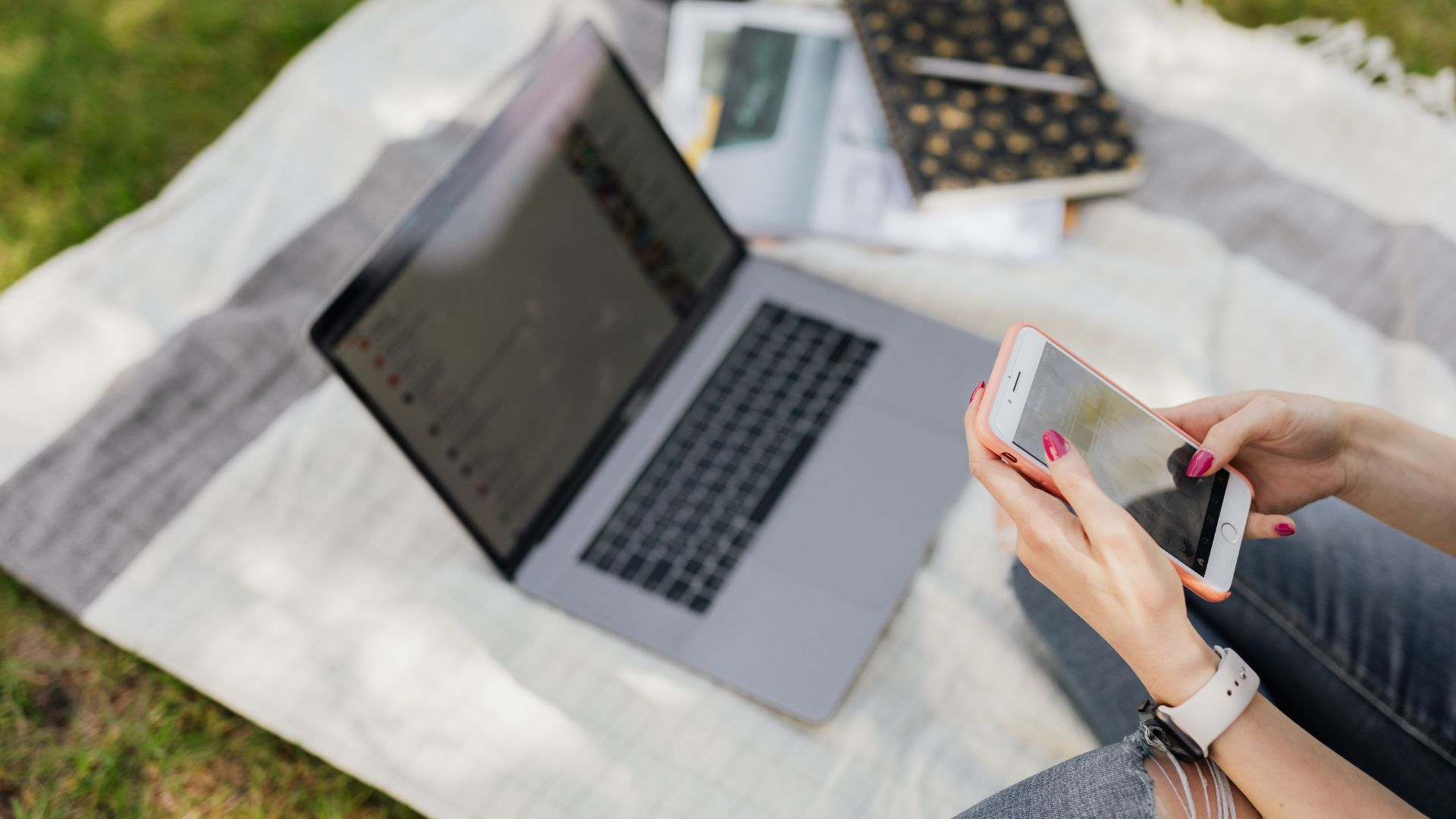 A woman is using a smartphone to ask a question to an AI or search engine while her laptop is open, working or researching outdoors on the grass