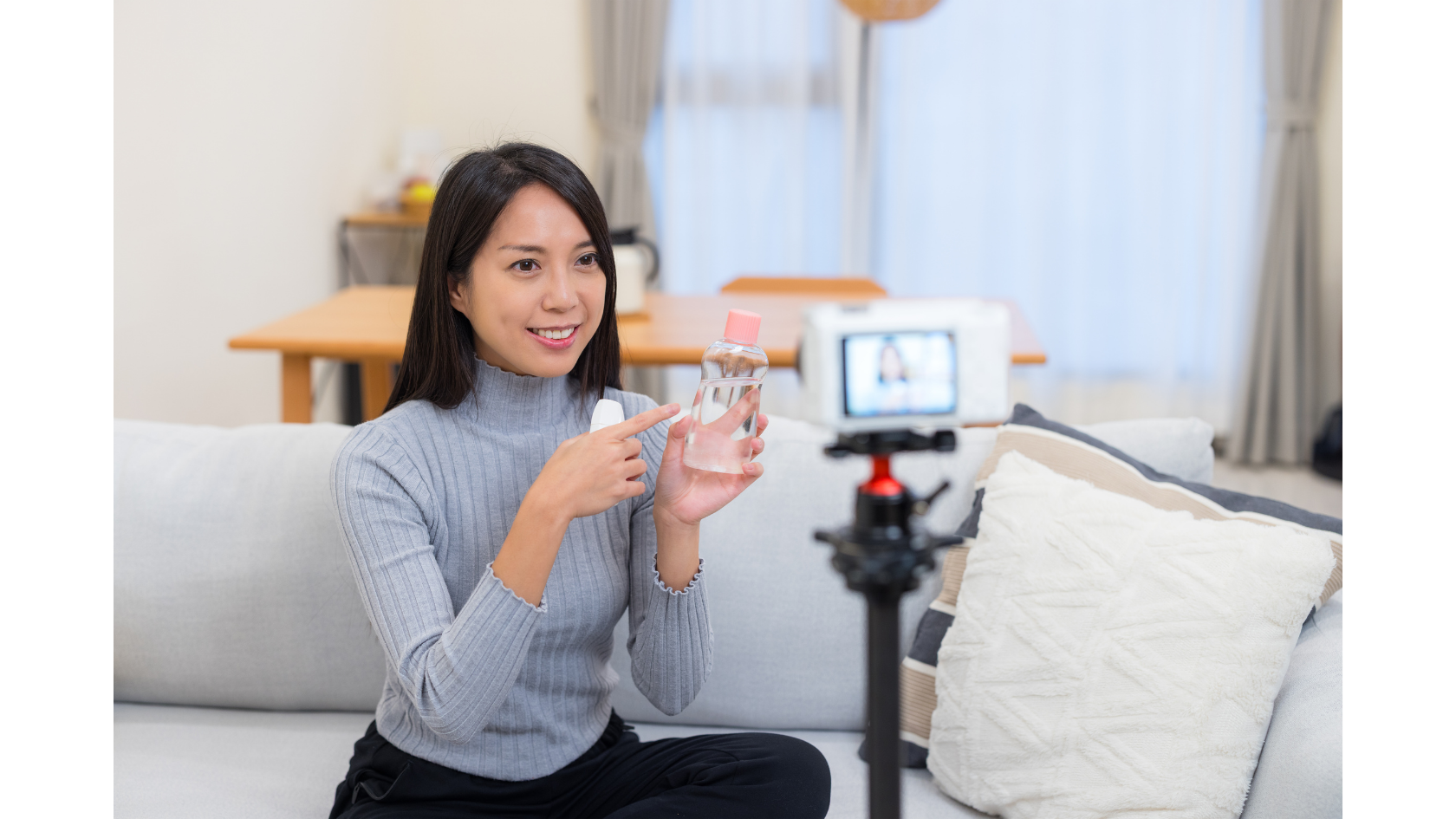 A female KOC (Key Opinion Consumer) recording a product review video at home, pointing at a beauty product in front of a camera tripod.