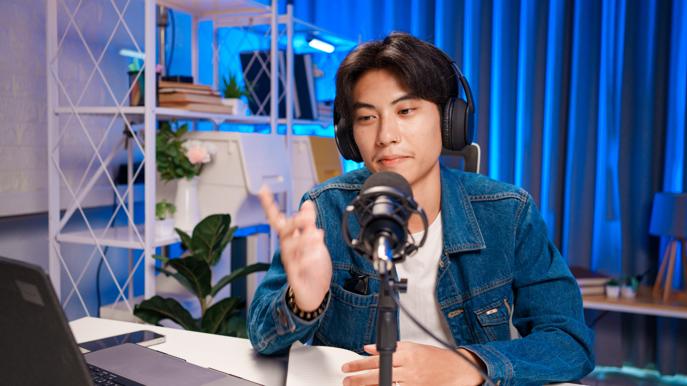 A young Asian man wearing a denim jacket and over-ear headphones sits at a white desk, speaking into a professional microphone. He is actively gesturing with his hand while looking toward a laptop screen, illustrating a live-streaming or real-time podcasting setup. The background features a well-organized room with a decorative shelf and vibrant blue ambient lighting, reflecting current high-engagement digital content trends.