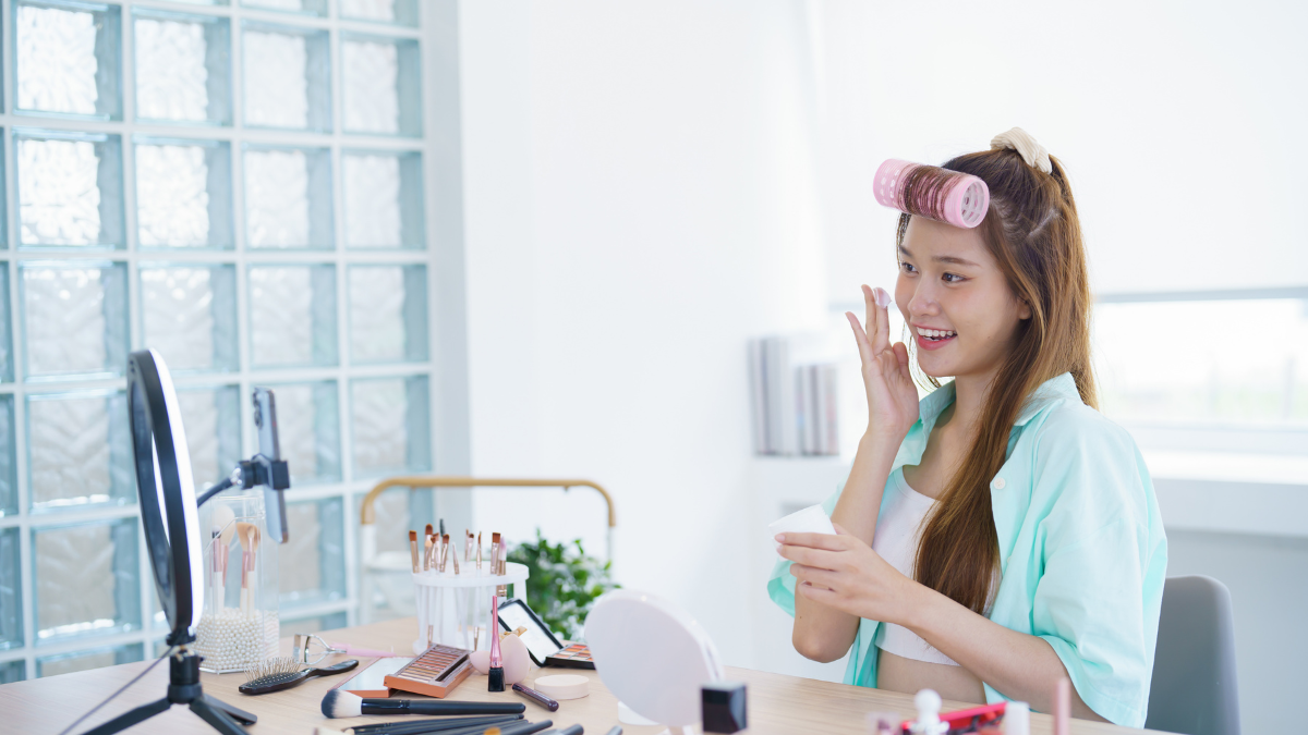 A beauty influencer with hair rollers applies facial cream while recording a makeup tutorial with a ring light and phone in her room.