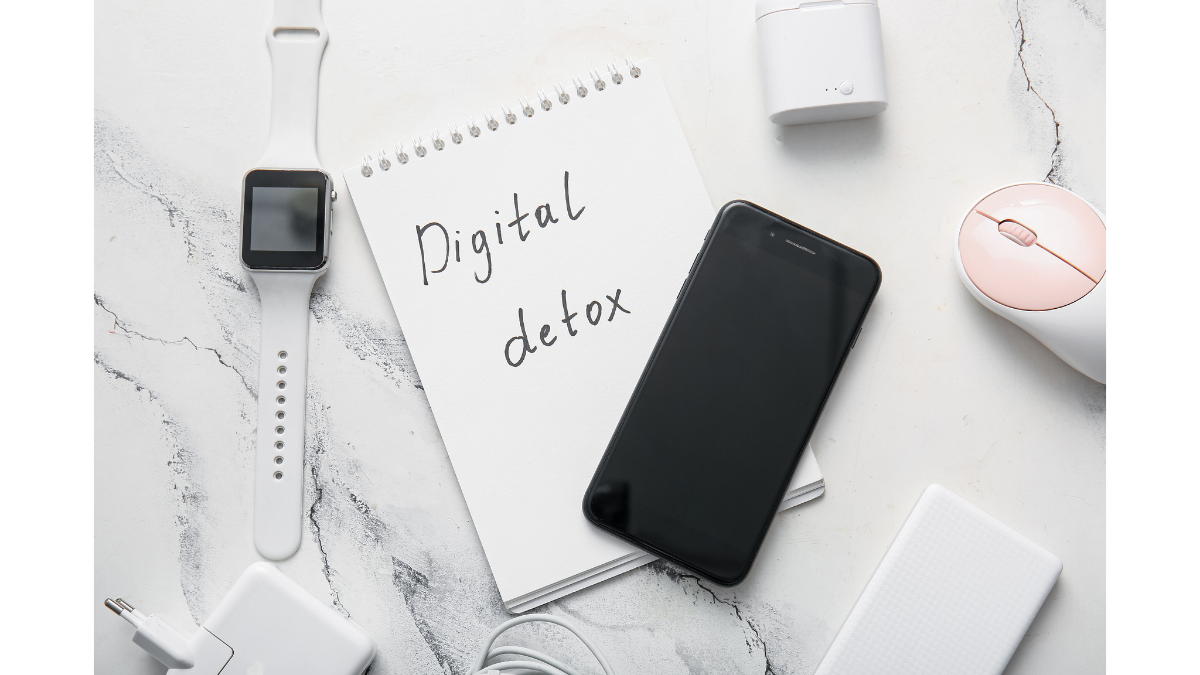 A top-down view of a white marble surface featuring a spiral notebook with the handwritten words "Digital detox." The notebook is surrounded by various tech gadgets, including a smartphone, a smartwatch, a computer mouse, and charging cables, all neatly arranged but pushed aside to focus on the message.