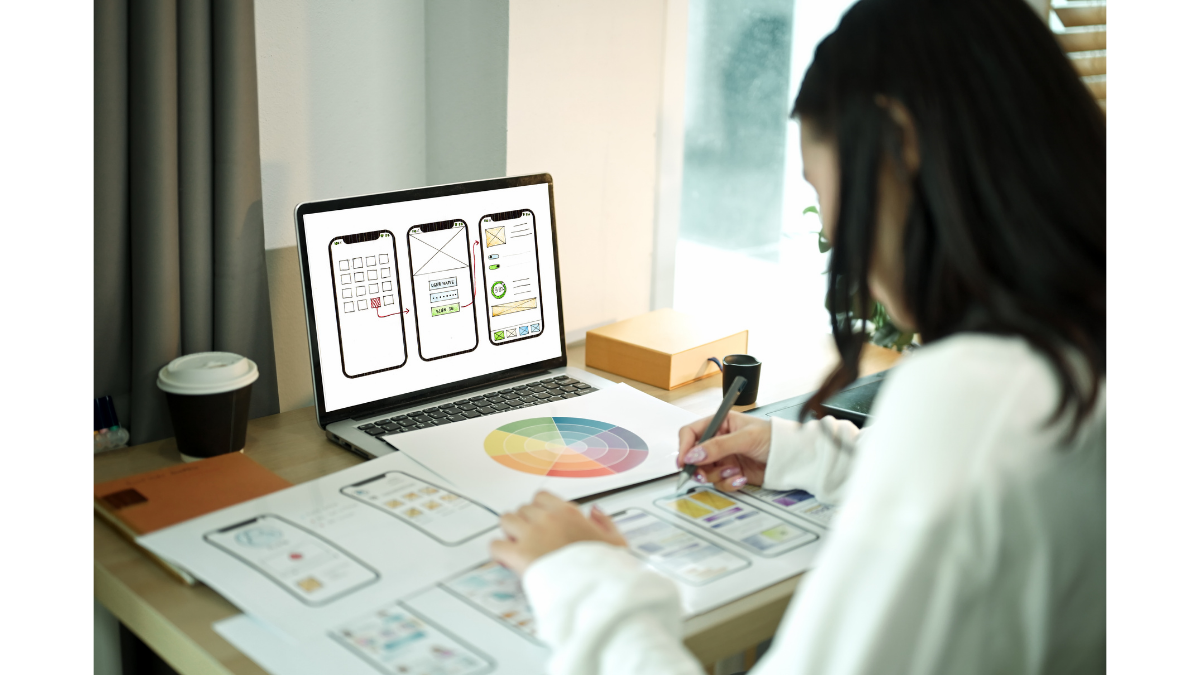 A female designer working at a desk, sketching mobile app wireframes on paper while a laptop in front of her displays digital UI mockups. A color wheel chart and a cup of coffee are also on the desk, illustrating the creative UX/UI design process.