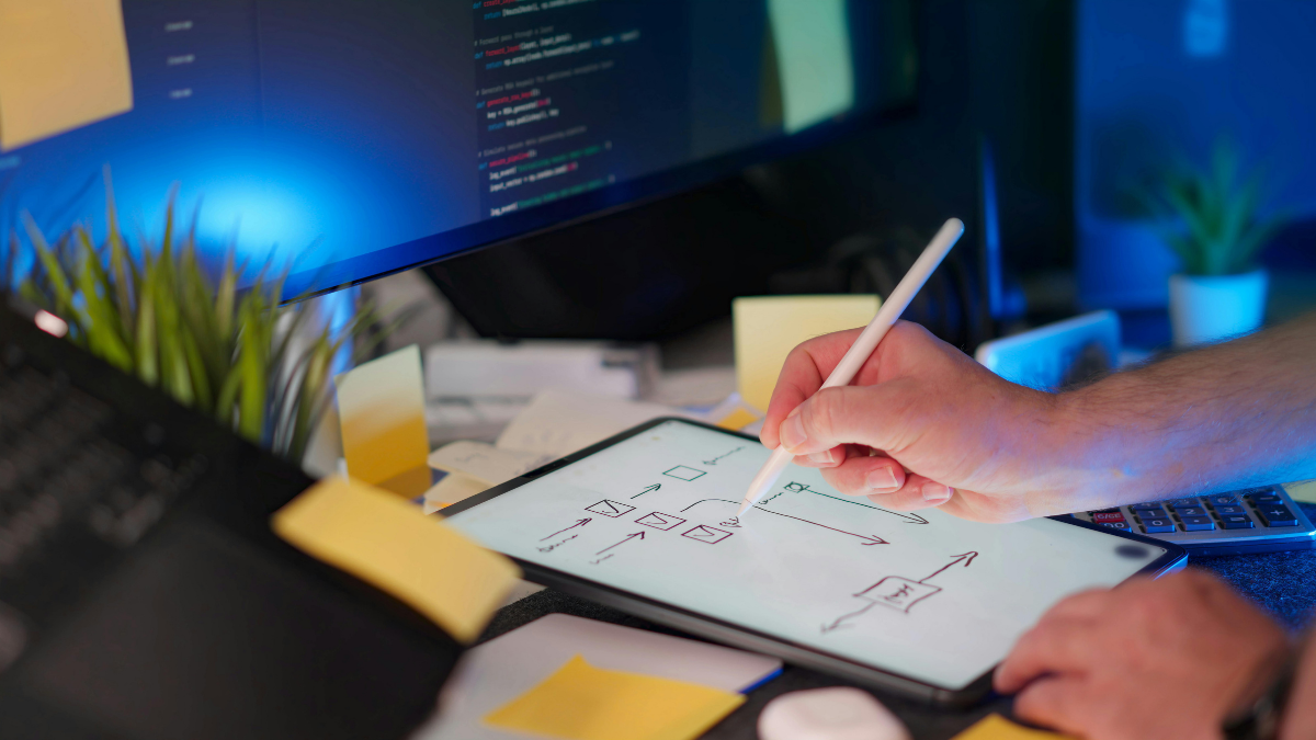 Digital ideation process: A UI designer mapping out a system architecture or user journey on a tablet, surrounded by sticky notes and a computer screen showing backend programming code.