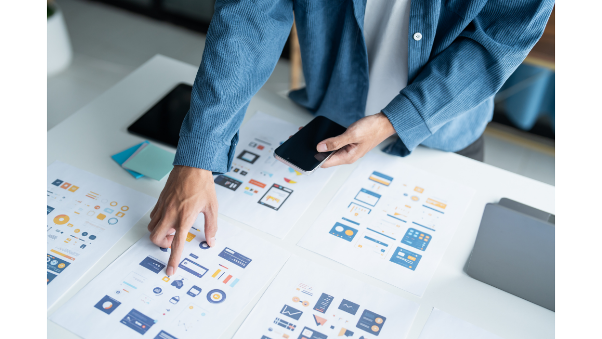 A designer sketching mobile app wireframes on paper while a laptop displays matching digital interface concepts, with a color wheel and coffee cup on the desk.