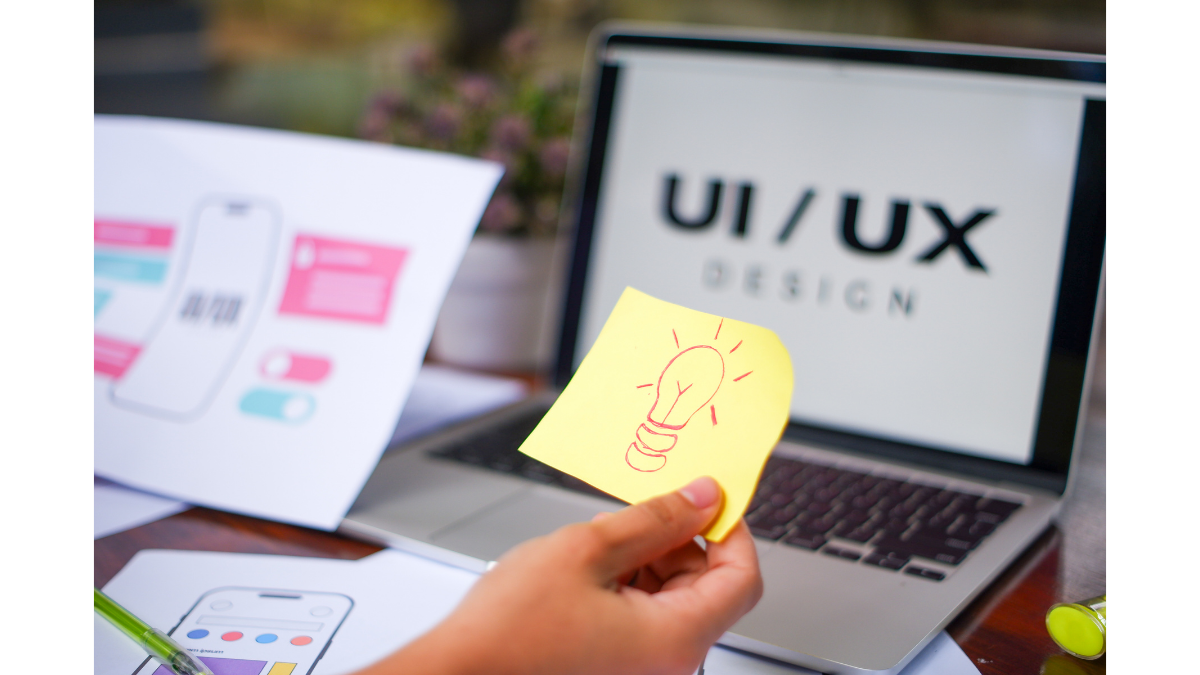 A designer's workspace featuring a laptop screen displaying 'UI/UX DESIGN,' hand-drawn mobile wireframes on paper, and a hand holding a yellow sticky note with a lightbulb icon, symbolizing creative brainstorming for design trends.