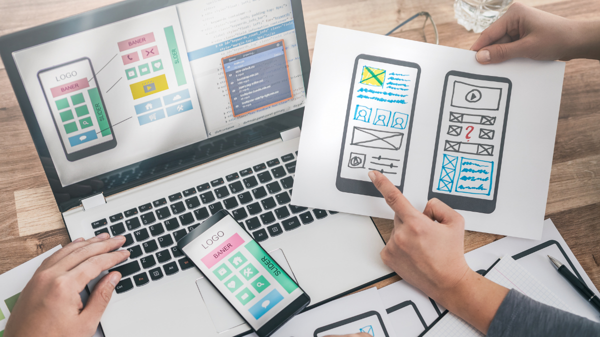An overhead view of a design team collaborating on user experience (UX) and user interface (UI). One hand points to detailed, hand-drawn wireframes for a mobile interface on a piece of paper. Next to it, a laptop screen displays code on one side and a corresponding digital design mockup on the other. A smartphone showing the same digital mockup is placed on the keyboard. They are meticulously planning the user journey to optimize the layout and features, with the goal of ultimately driving more website sales.