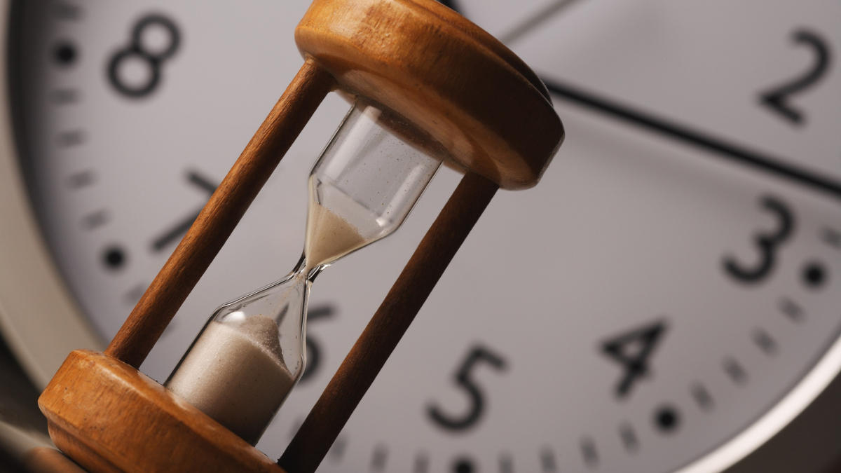 Close-up of a classic wooden hourglass with sand flowing, set against the blurred face of a modern analog clock. Therefore, this image illustrates the crucial concept of timing and speed required for successful, high-engagement real-time marketing.
