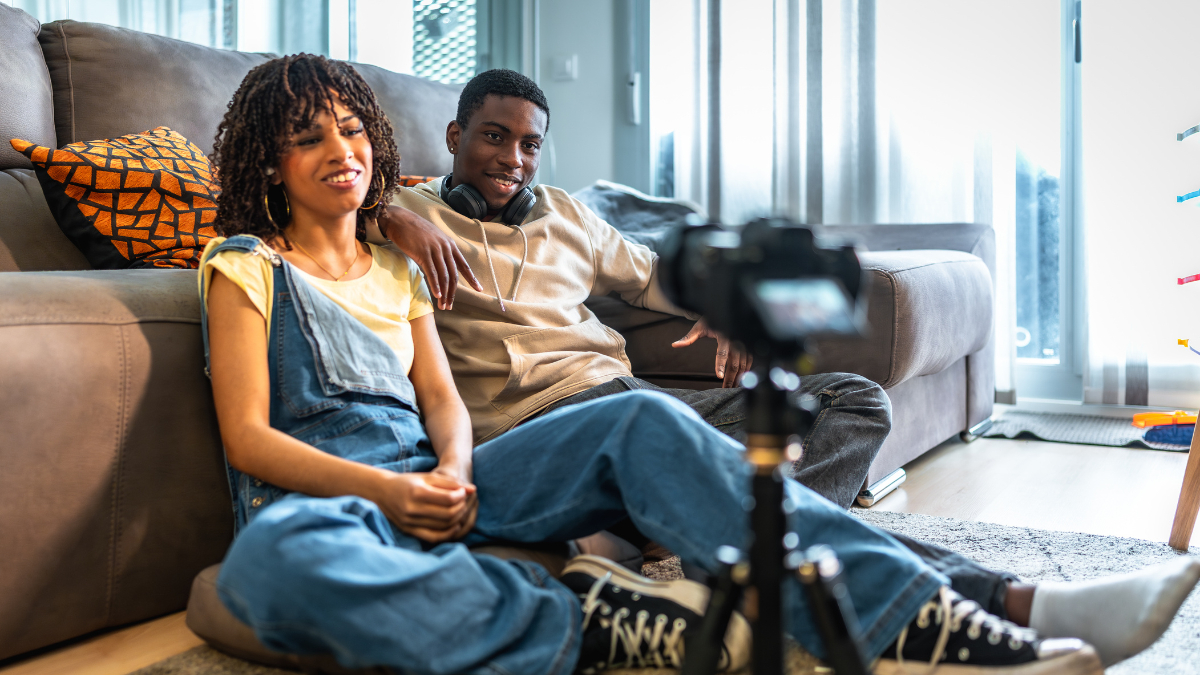 Two young digital creators sitting on a living room floor, smiling and recording a live video or vlog on a camera. Therefore, this image exemplifies the authentic, raw, and relatable nature of user-generated content that drives high-engagement trends today.
