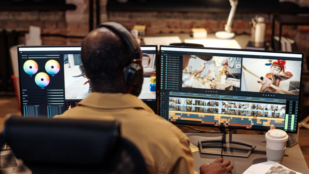 View from behind of a professional video editor working diligently at a dual-monitor workstation. The left screen displays color grading tools, while the right screen shows a video editing timeline. Therefore, this image represents the rapid, high-quality content creation essential for capturing high-engagement digital trends.