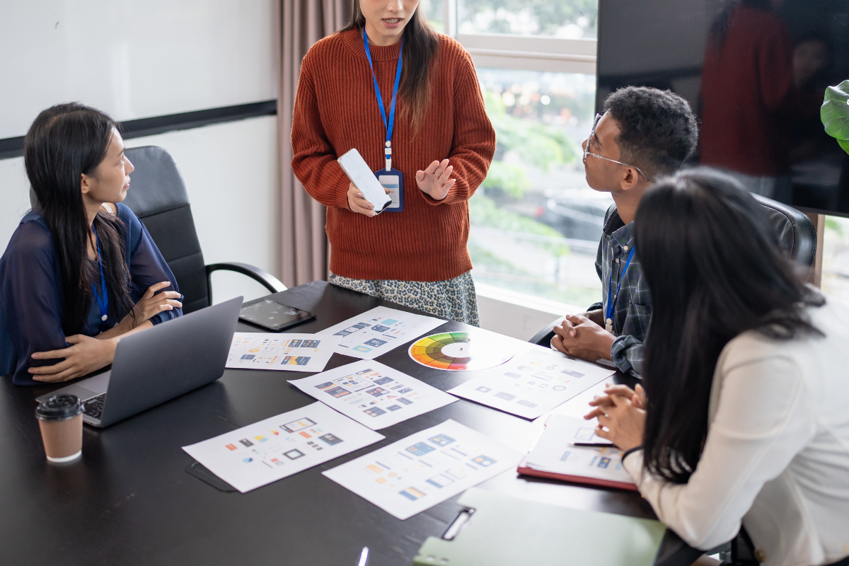 A creative team brainstorming social media content trends. They are reviewing mobile app mockups and a color wheel spread across a conference table.
