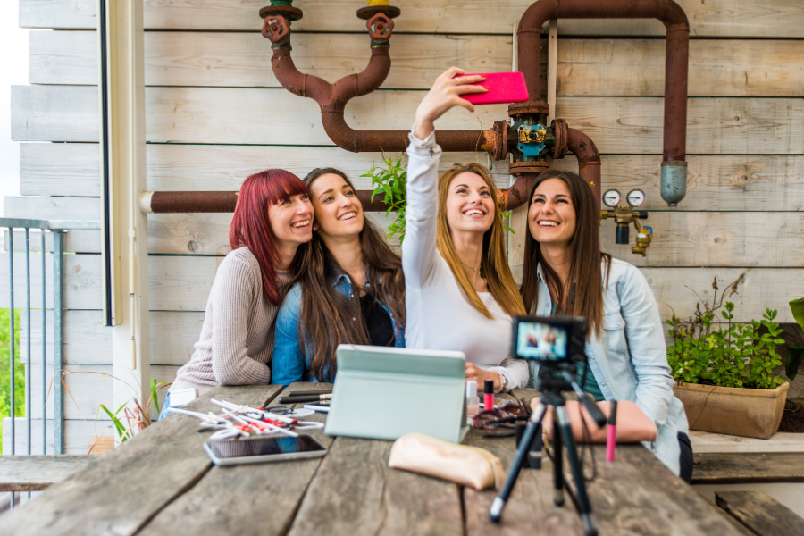Four female content creators smiling and taking a selfie together at a wooden table with a camera, makeup products, and a tablet, representing Influencer marketing and social media content creation in a casual lifestyle setting.