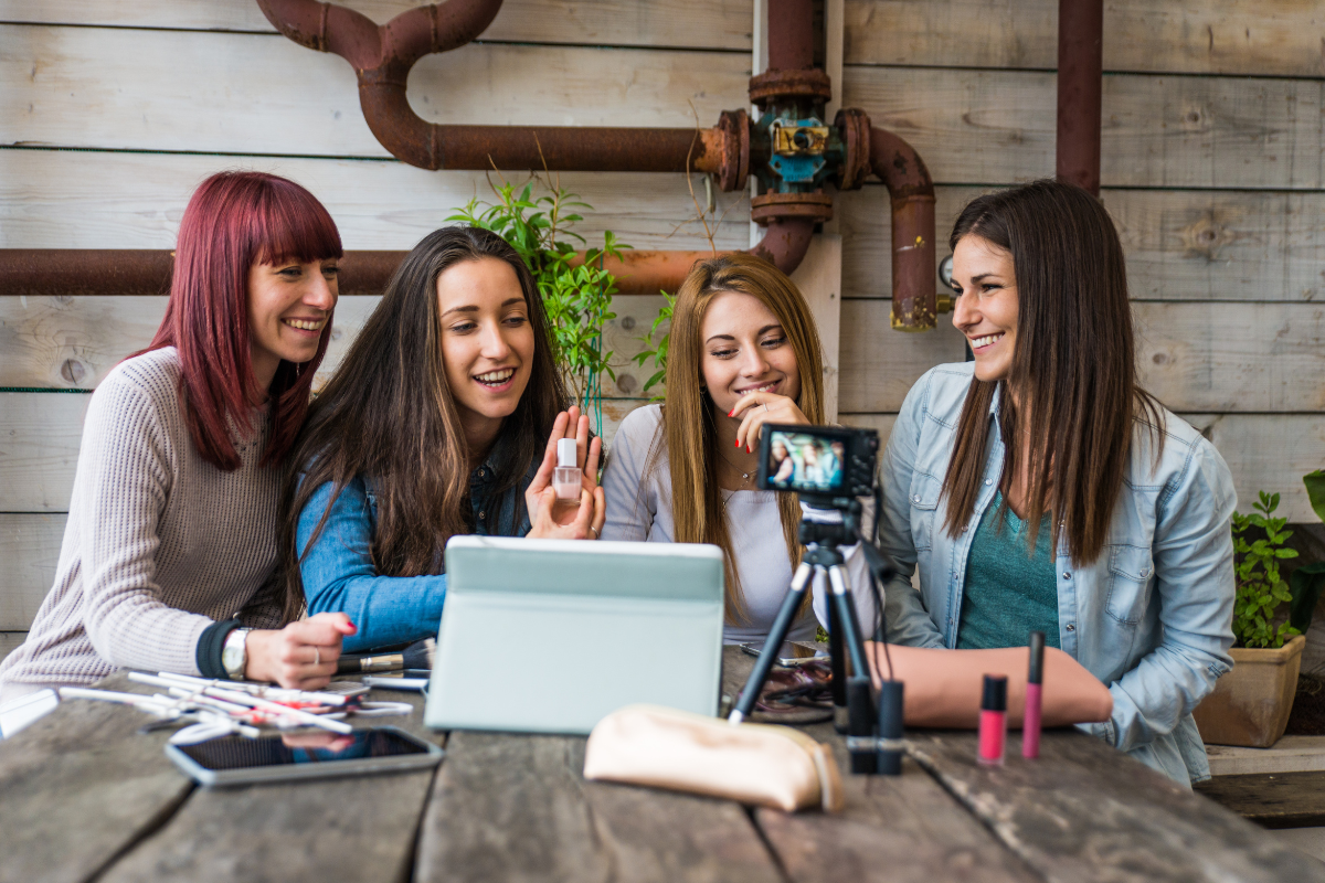 Four female content creators smiling and taking a selfie together at a wooden table with a camera, makeup products, and a tablet, representing Influencer marketing and social media content creation in a casual lifestyle setting.