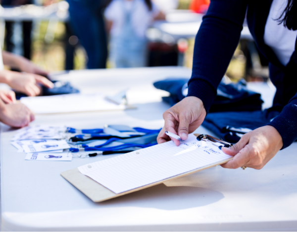 A close-up of a person signing a manual paper registration sheet on a clipboard, representing a traditional check-in method compared to modern event registration kiosks.