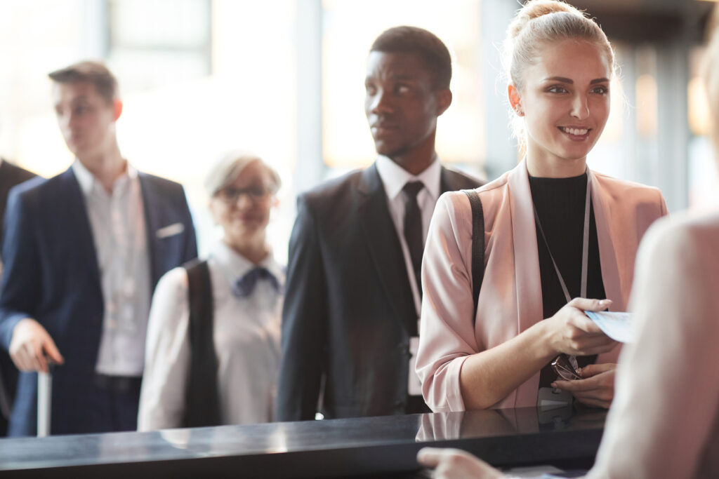 business-professionals-standing-in-line-at-an-event-registration-desk-with-a-smiling-woman-in-a-pink-blazer-checking-in-at-the-counter