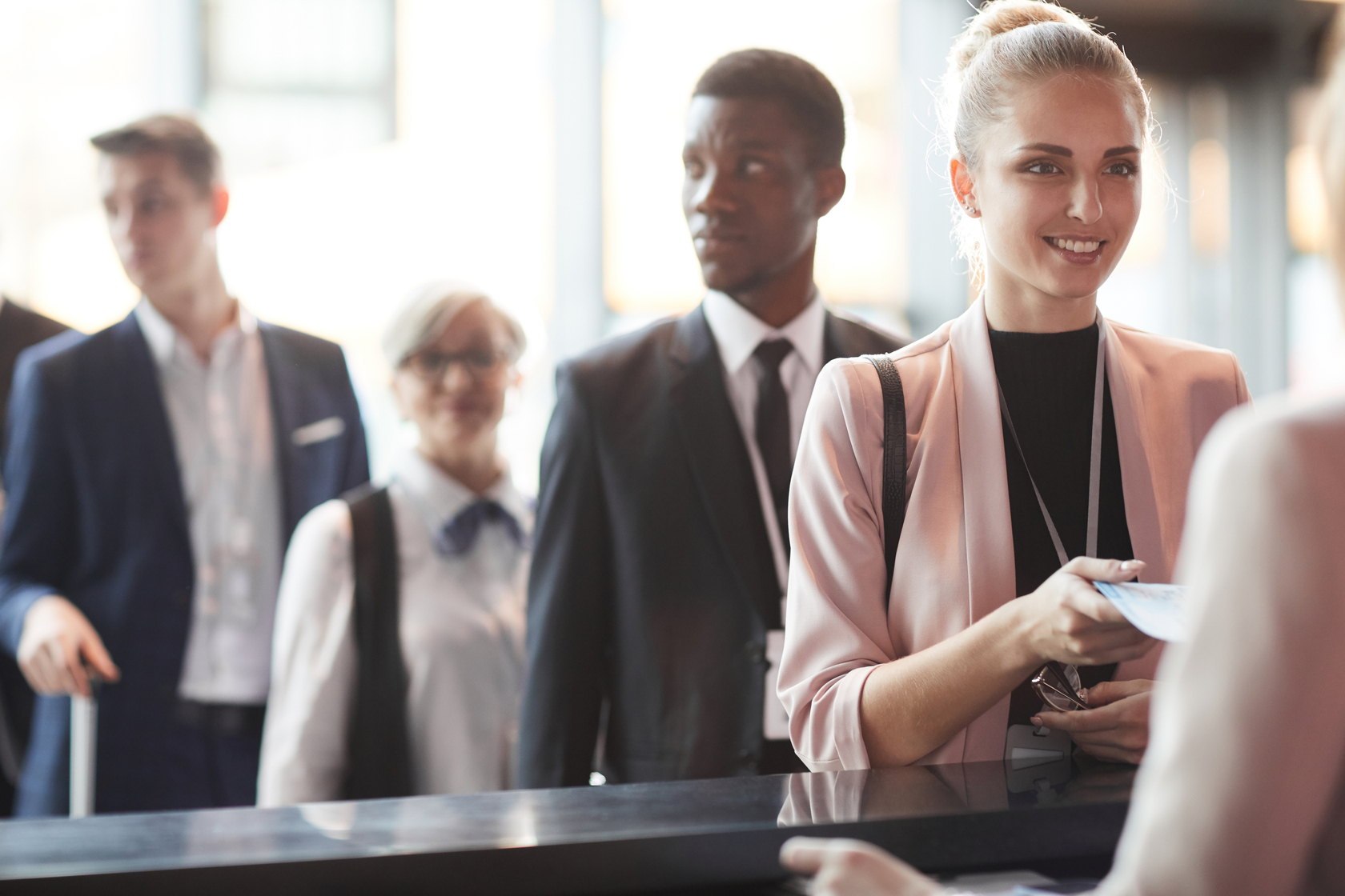 business-professionals-standing-in-line-at-an-event-registration-desk-with-a-smiling-woman-in-a-pink-blazer-checking-in-at-the-counter